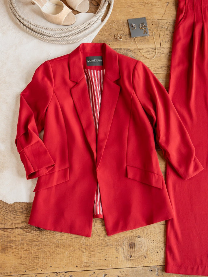 The Revillo Red Blazer Set by Savannah Sevens western life{&}style, featuring a red satin blazer with striped lining, is laid flat on a wooden surface alongside the matching high waisted pants, beige sandals, and gold jewelry.