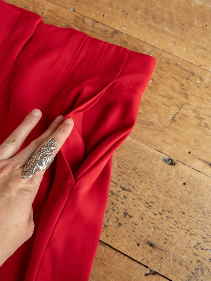 A hand adorned with a large silver ring holds open a pleat on the high-waisted pants from the Revillo Red Blazer Set by Savannah Sevens western life{&}style, displayed on a wooden surface.
