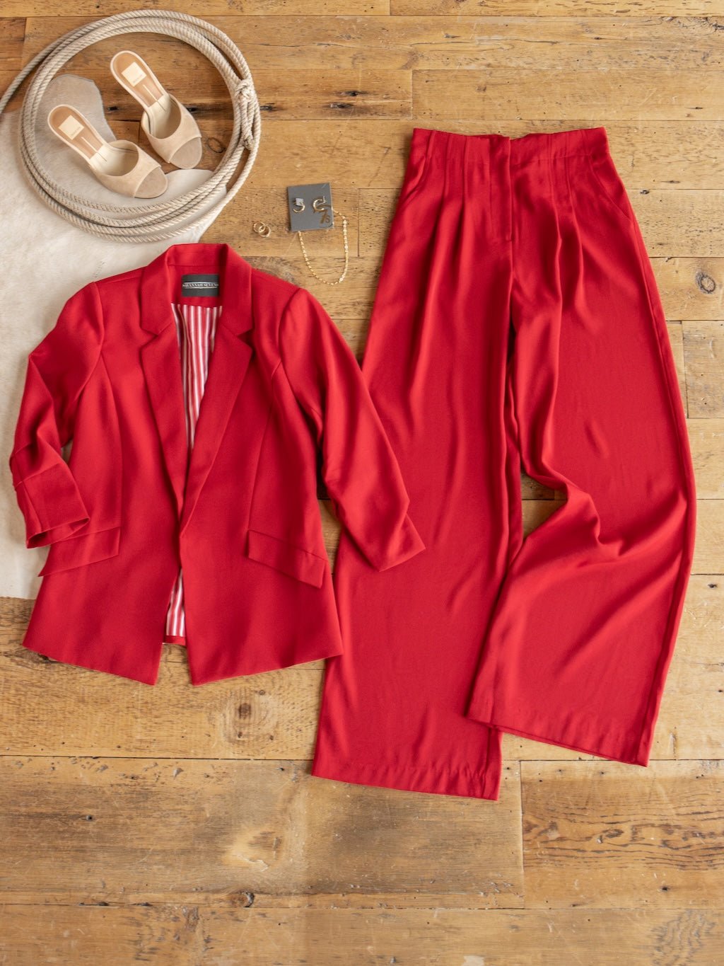The Revillo Red Blazer Set by Savannah Sevens western life{&}style, featuring wide-leg satin trousers and a coordinating blazer, is displayed on a wooden floor alongside beige sandals and a necklace.