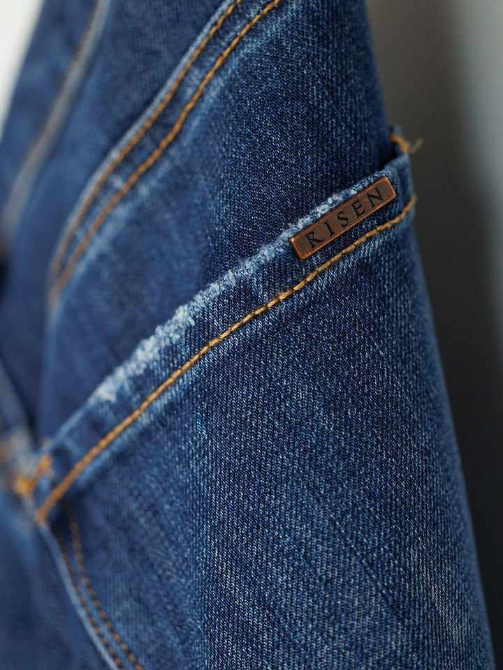Close-up of blue denim The Rhett High Rise Relaxed Straight Leg Jean by Savannah Sevens western life{&}style, featuring orange stitching and a small metallic tag near the seam. These jeans highlight quality craftsmanship and classic style.
