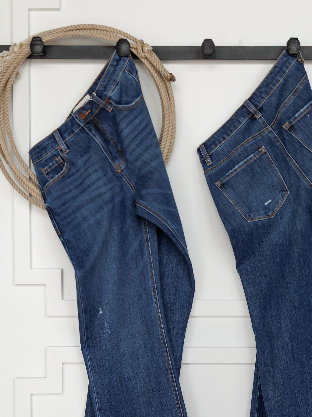 Two pairs of The Rhett High Rise Relaxed Straight Leg Jeans by Savannah Sevens western life{&}style hang from black hooks on a white wall, with a coiled rope beside them for a rustic, western accent in the minimalist space.