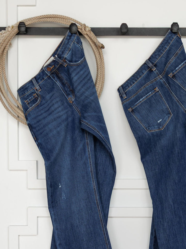 Two pairs of The Rhett High Rise Relaxed Straight Leg Jeans by Savannah Sevens western life{&}style hang from black hooks on a white wall, with a coiled rope beside them for a rustic, western accent in the minimalist space.