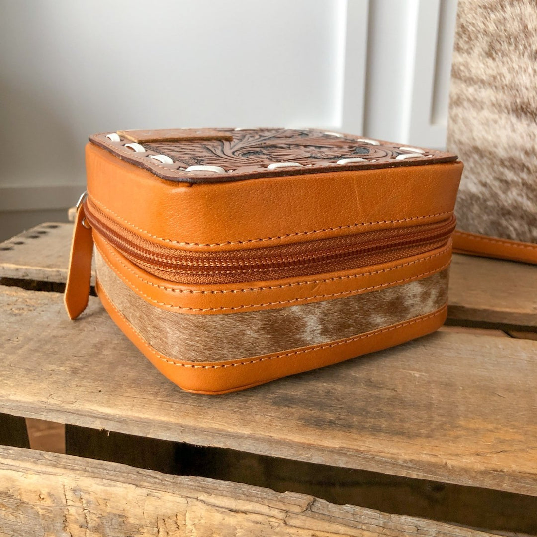 The Rialto Tooled Leather Mini Travel Jewelry Case by Savannah Sevens western life{&}style is a small, square brown cowhide case with a zipper and tooled leather detail, shown on a rustic wooden surface.