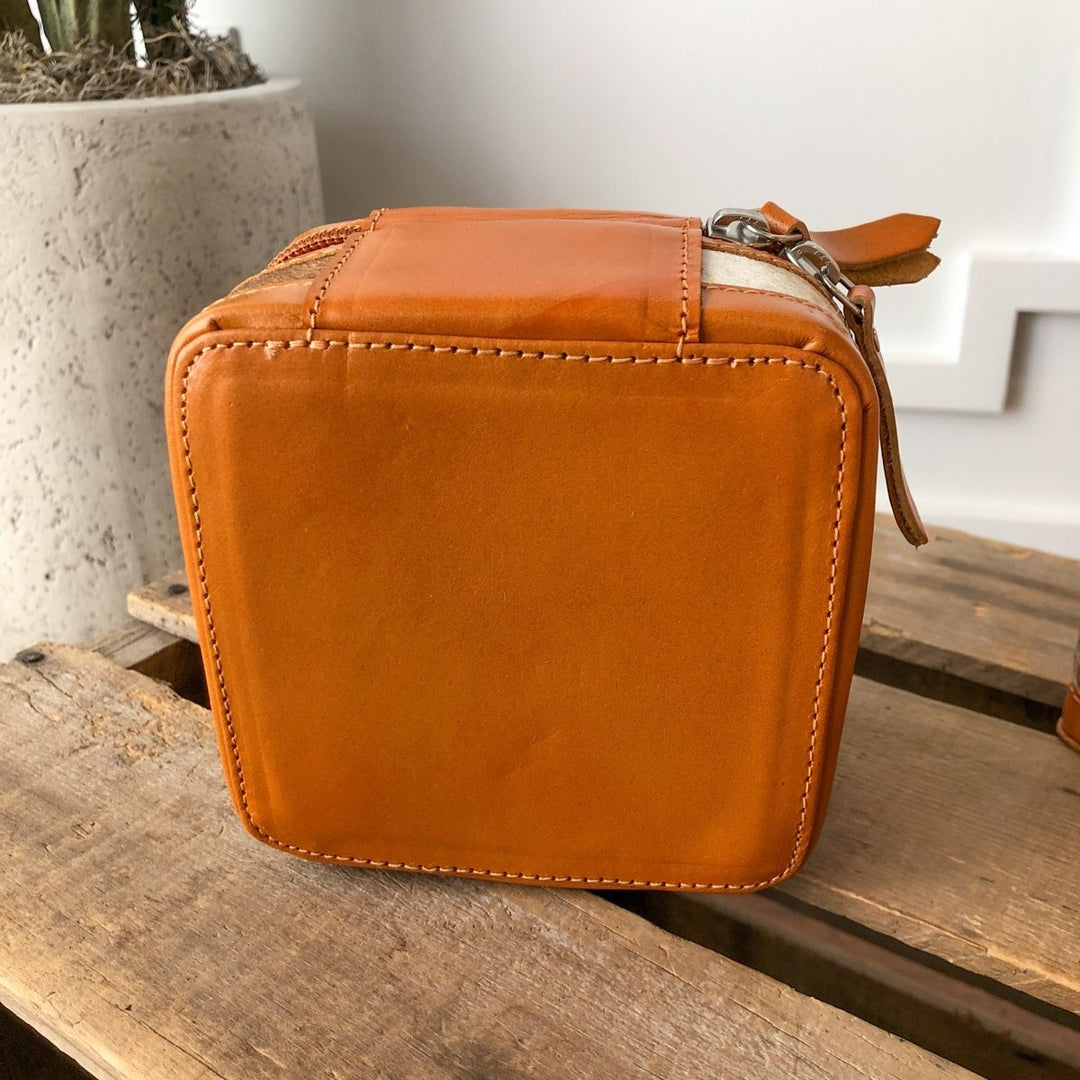 The Rialto Tooled Leather Mini Travel Jewelry Case by Savannah Sevens western life{&}style, a small square orange cowhide case with zipper and strap, sits on rustic wood beside a textured concrete planter.
