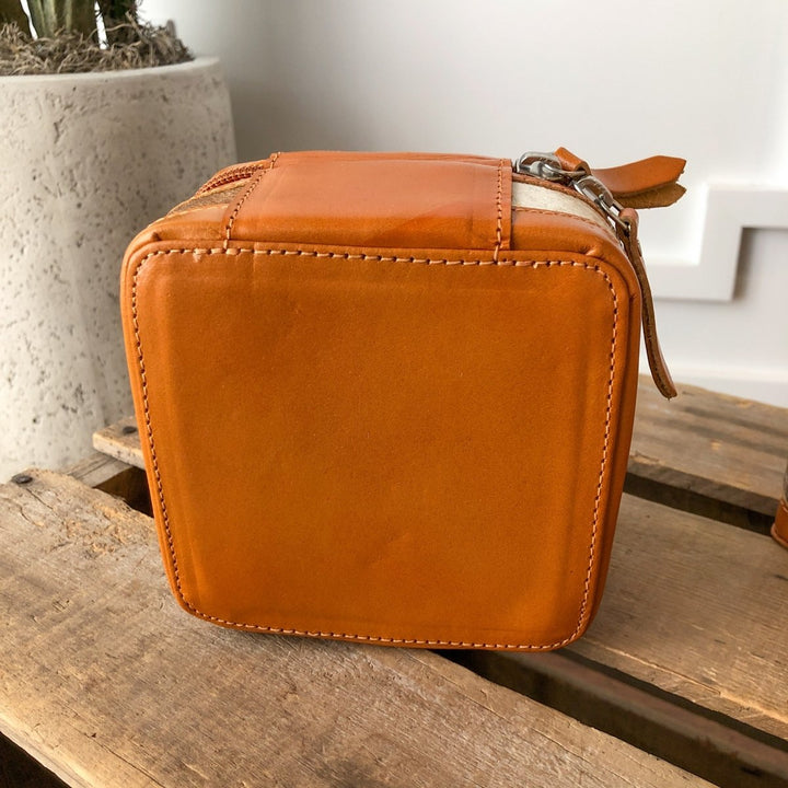 The Rialto Tooled Leather Mini Travel Jewelry Case by Savannah Sevens western life{&}style, a small square orange cowhide case with zipper and strap, sits on rustic wood beside a textured concrete planter.