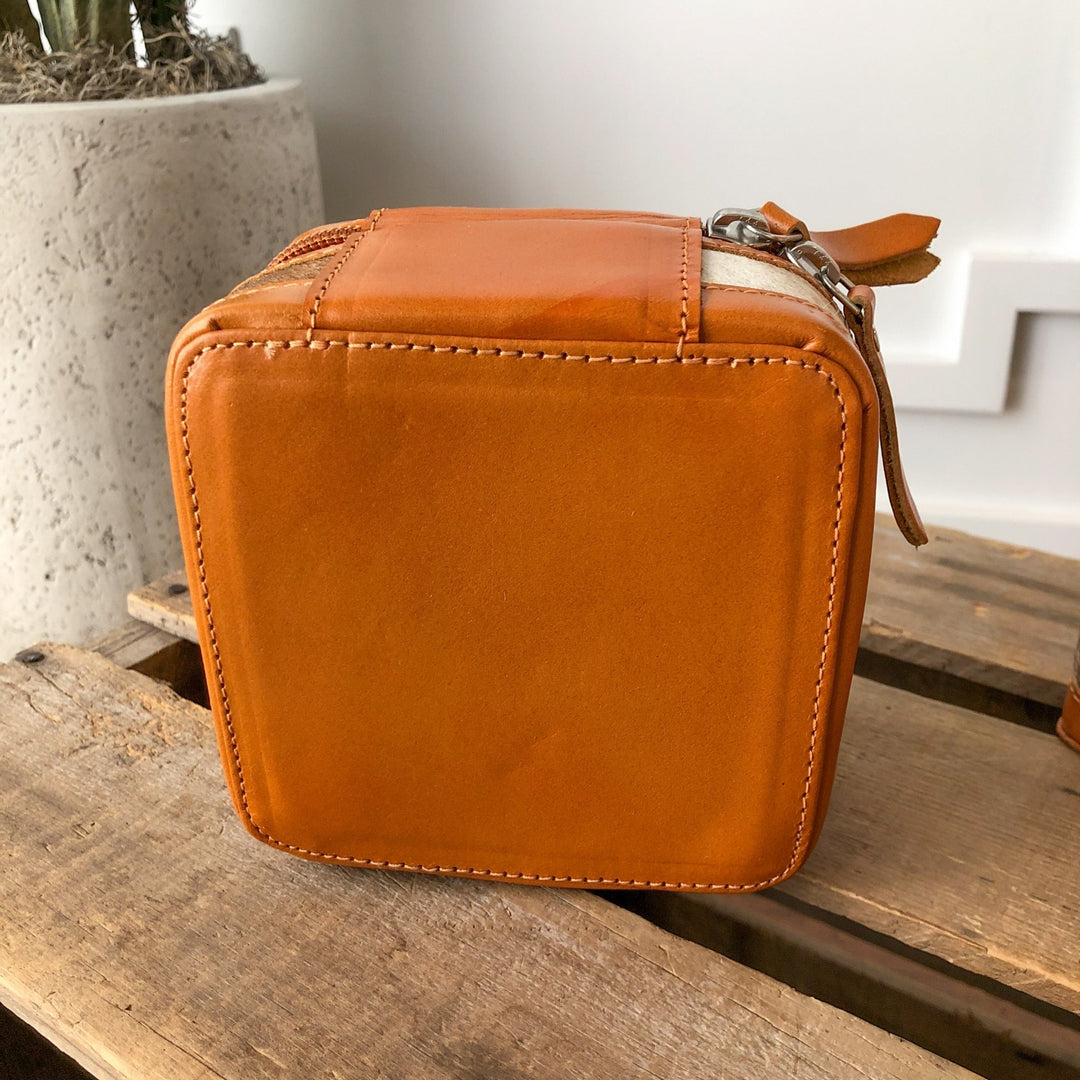 The Rialto Tooled Leather Mini Travel Jewelry Case by Savannah Sevens western life{&}style, a small square orange cowhide case with zipper and strap, sits on rustic wood beside a textured concrete planter.