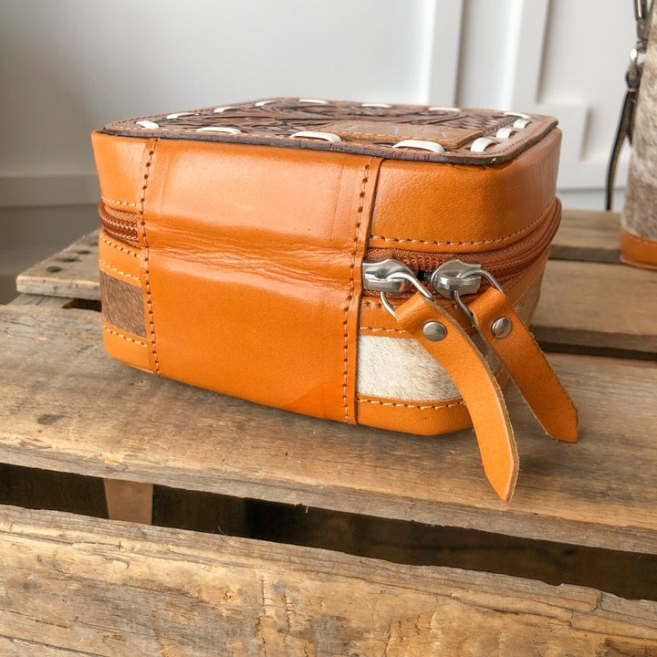 The Rialto Tooled Leather Mini Travel Jewelry Case by Savannah Sevens western life{&}style is a small, square brown leather pouch with white stitching, decorative patterns, two zipper pulls, and light/dark leather accents on a rustic wooden surface.