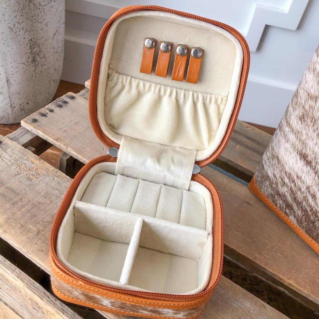 The Rialto Tooled Leather Mini Travel Jewelry Case by Savannah Sevens western life{&}style features a tan and cream interior with ring slots, divided compartments, and lid holders for earrings or necklaces. It's pictured resting on a wooden surface.