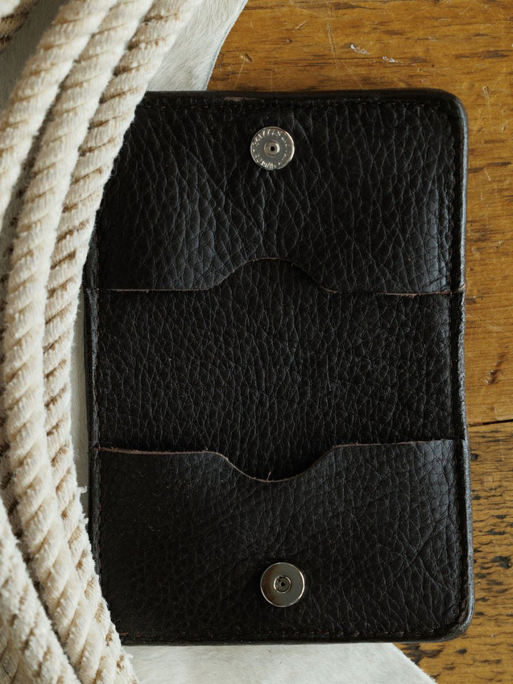 A black leather Rialto Wallet with boot stitch detail and two snap buttons lies open on a wooden surface, framed by coiled rope and soft white fabric. Crafted by Savannah Sevens western life{&}style.
