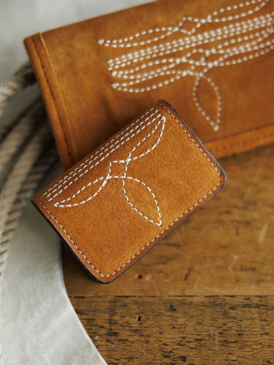 The Rialto Wallet by Savannah Sevens western life{&}style, a small brown leather card wallet with white boot stitch detail, rests on wood beside a matching larger Double J Saddlery item, both showcasing similar stitching and a coiled rope.