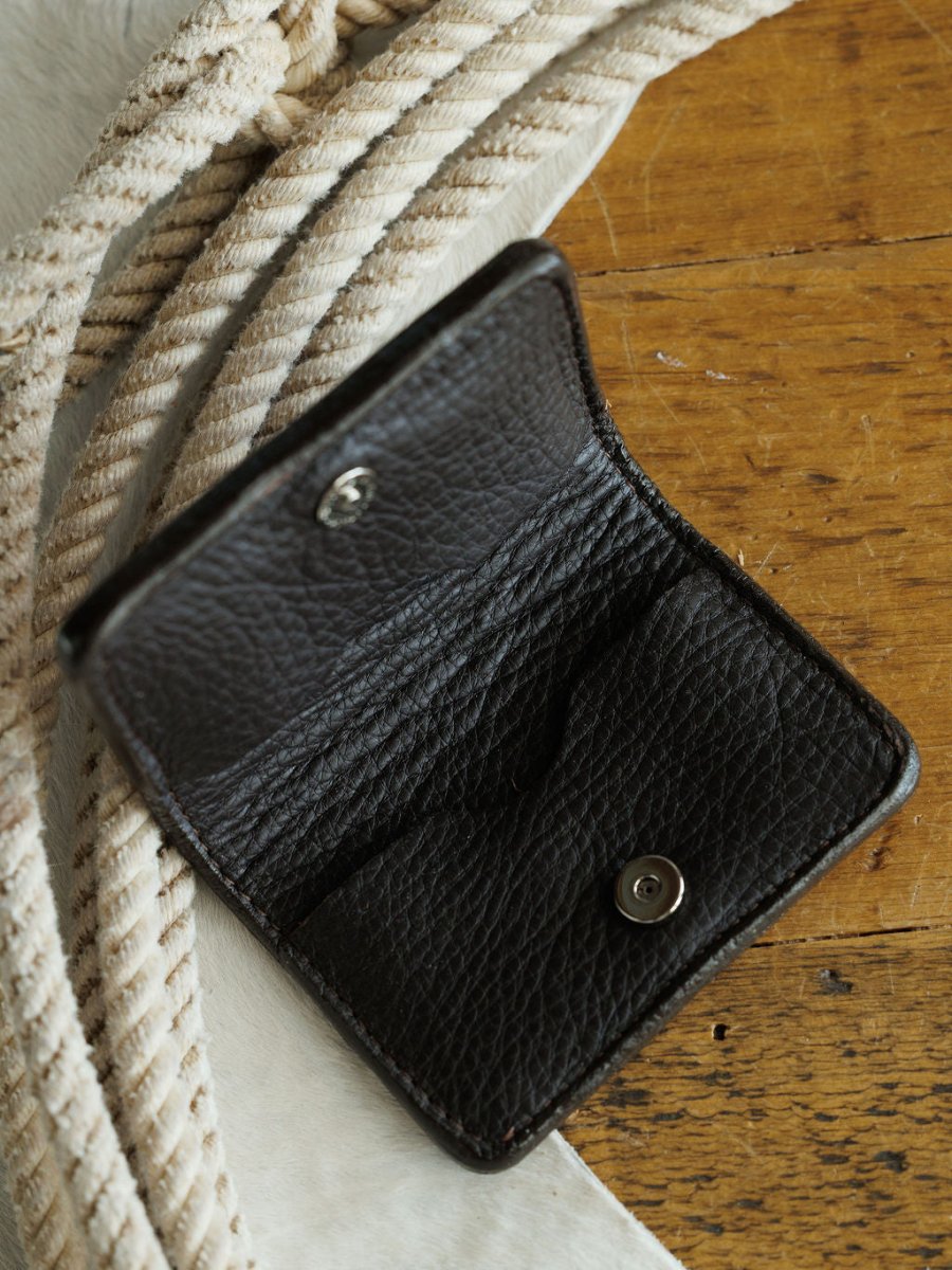 The Rialto Wallet by Savannah Sevens western life{&}style, featuring open textured black leather with a snap button and boot stitch detail, rests on a wooden surface beside coiled white rope.