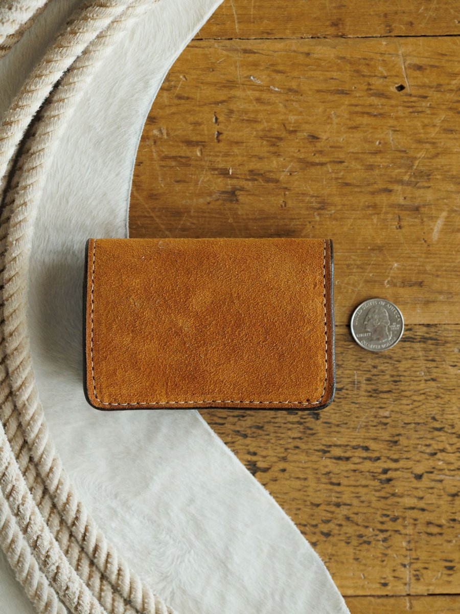 The Rialto Wallet by Savannah Sevens western life{&}style, a small brown leather card wallet with boot stitch detail, rests on a wooden surface next to a U.S. quarter, with white rope and fabric visible in the corner.