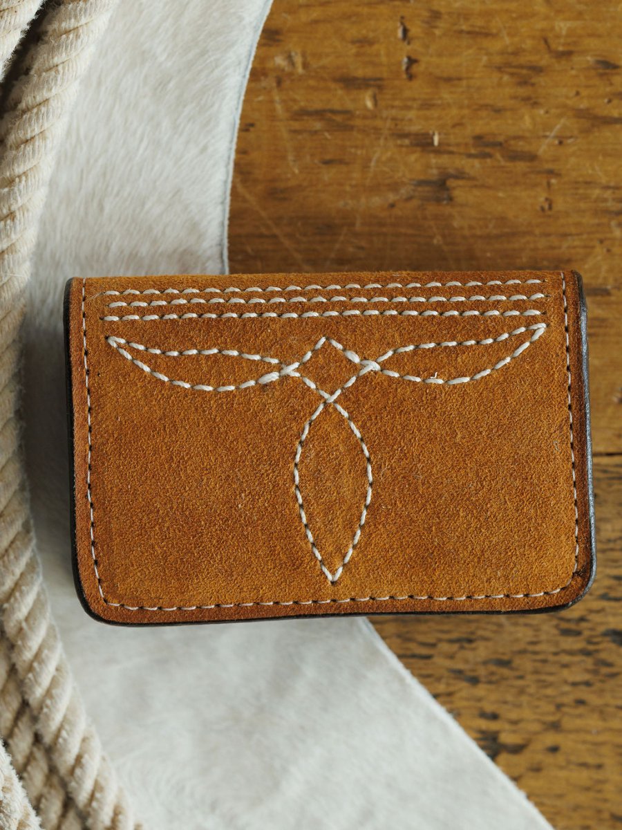 The Rialto Wallet by Savannah Sevens western life{&}style, a small brown leather card wallet with white boot stitch detail and a symmetrical pattern, rests on wood beside light cowhide and textured beige fabric—crafted with Double J Saddlery artistry.