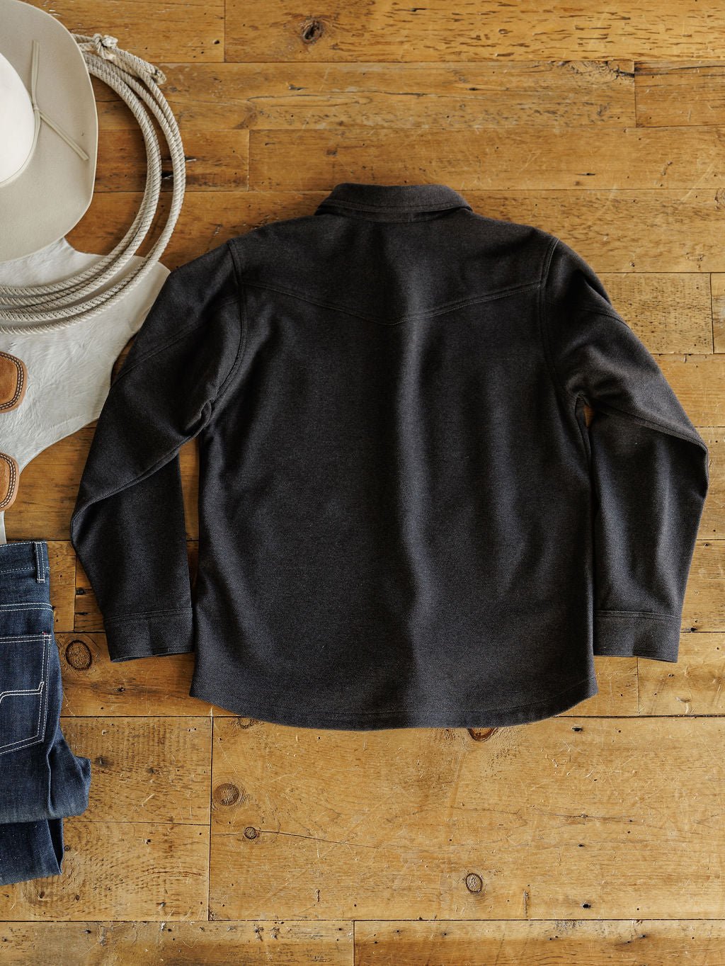 A dark long-sleeve shirt is laid flat on wood next to the Richford Ariat Men's Charcoal Jacket by Savannah Sevens western life{&}style, with a white cowboy hat, gloves, rope, and folded blue jeans for a rustic Western vibe.