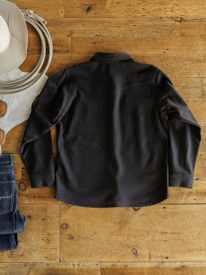 A dark long-sleeve shirt is laid flat on wood next to the Richford Ariat Men's Charcoal Jacket by Savannah Sevens western life{&}style, with a white cowboy hat, gloves, rope, and folded blue jeans for a rustic Western vibe.