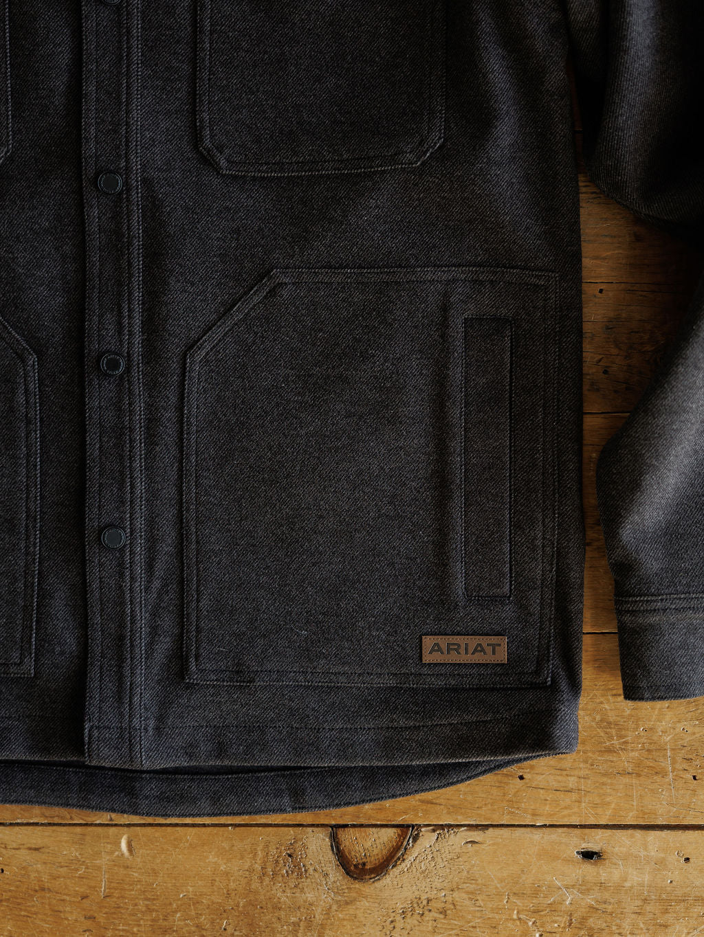 A close-up of the Richford Ariat Men's Charcoal Jacket by Savannah Sevens western life{&}style, laid flat on wood, featuring large front pockets, buttons, and a small brown Ariat label near the lower pocket.