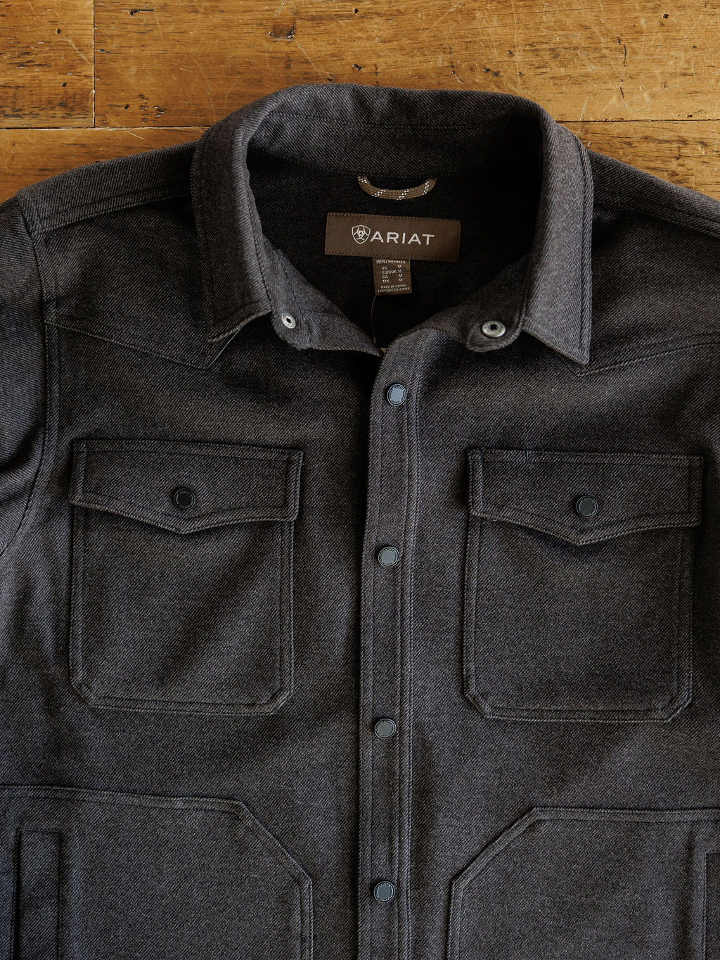 The Richford Ariat Men's Charcoal Jacket by Savannah Sevens western life{&}style, featuring snap buttons and two front chest pockets, lies flat on a wooden surface. This heather charcoal jacket is both stylish and functional for any occasion.
