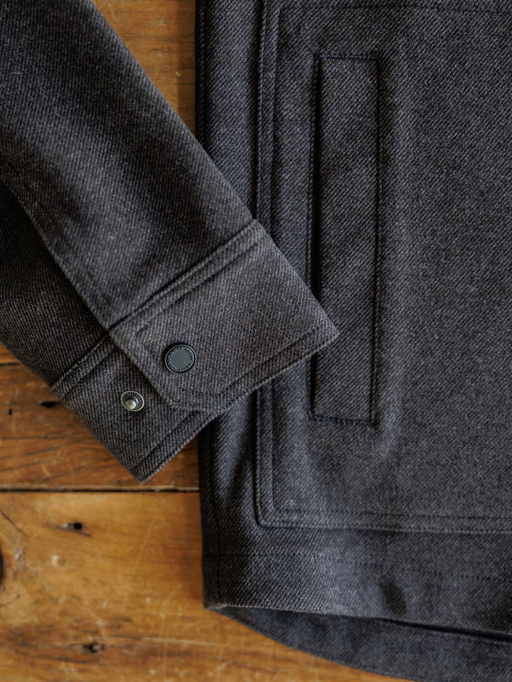 Close-up of a Richford Ariat Men's Charcoal Jacket sleeve by Savannah Sevens western life{&}style, featuring snap buttons and a front pocket, laid on a rustic wooden surface.