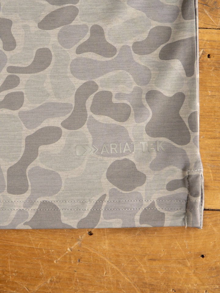 Close-up of the Ridgefield Ariat Men's Grey Camo Polo from Savannah Sevens western life{&}style, featuring gray camo fabric with "ARIAT TEK" print, moisture wicking, sun protection properties, and detailed hem stitching on a wooden surface.