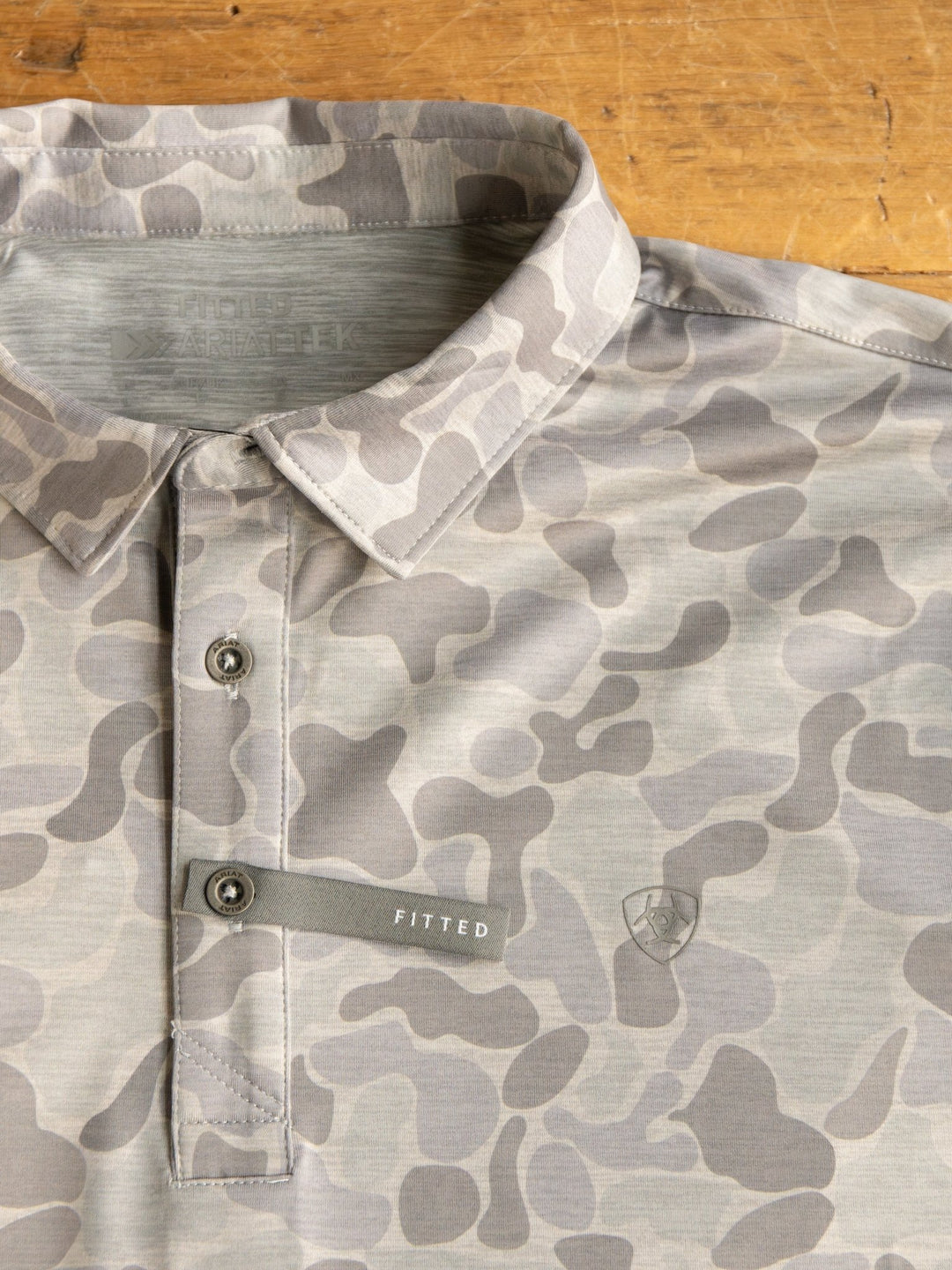The Ridgefield Ariat Men's Grey Camo Polo by Savannah Sevens western life{&}style features a button placket with a FITTED tag, moisture-wicking properties, and is shown on a wooden surface.