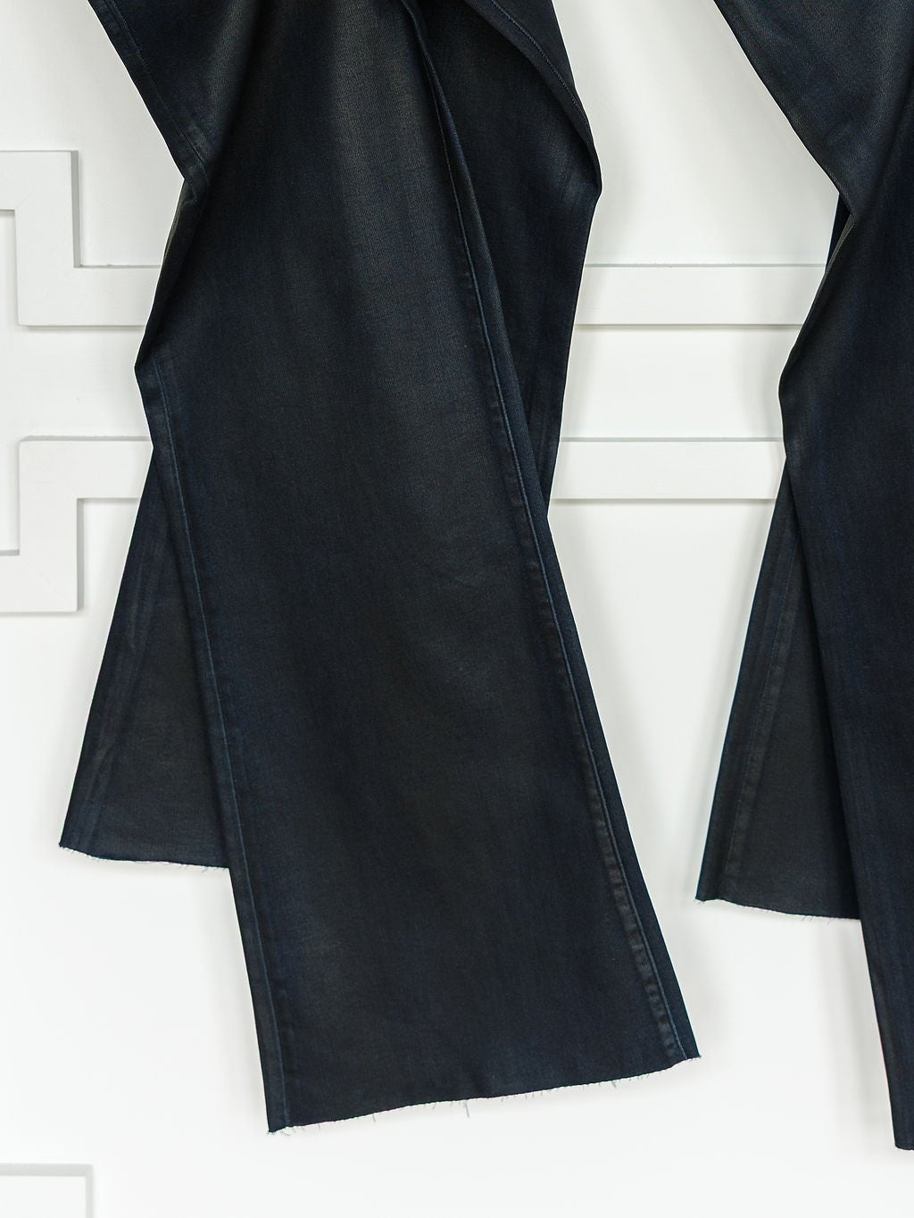 Close-up of The Riggins High Rise Wide Leg Jean by Savannah Sevens western life{&}style, featuring coated dark wash denim, a '90s wide leg silhouette, and raw unfinished hems, hanging against a white textured wall.