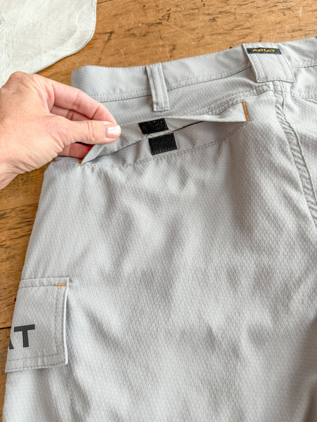 A hand lifts the flap of a light gray pair of Rockville Ariat Men's M5 Shorts by Savannah Sevens western life{&}style, showing a pocket with two black Velcro closures. The men's work shorts have textured fabric and a side pocket with CAT printed on it.