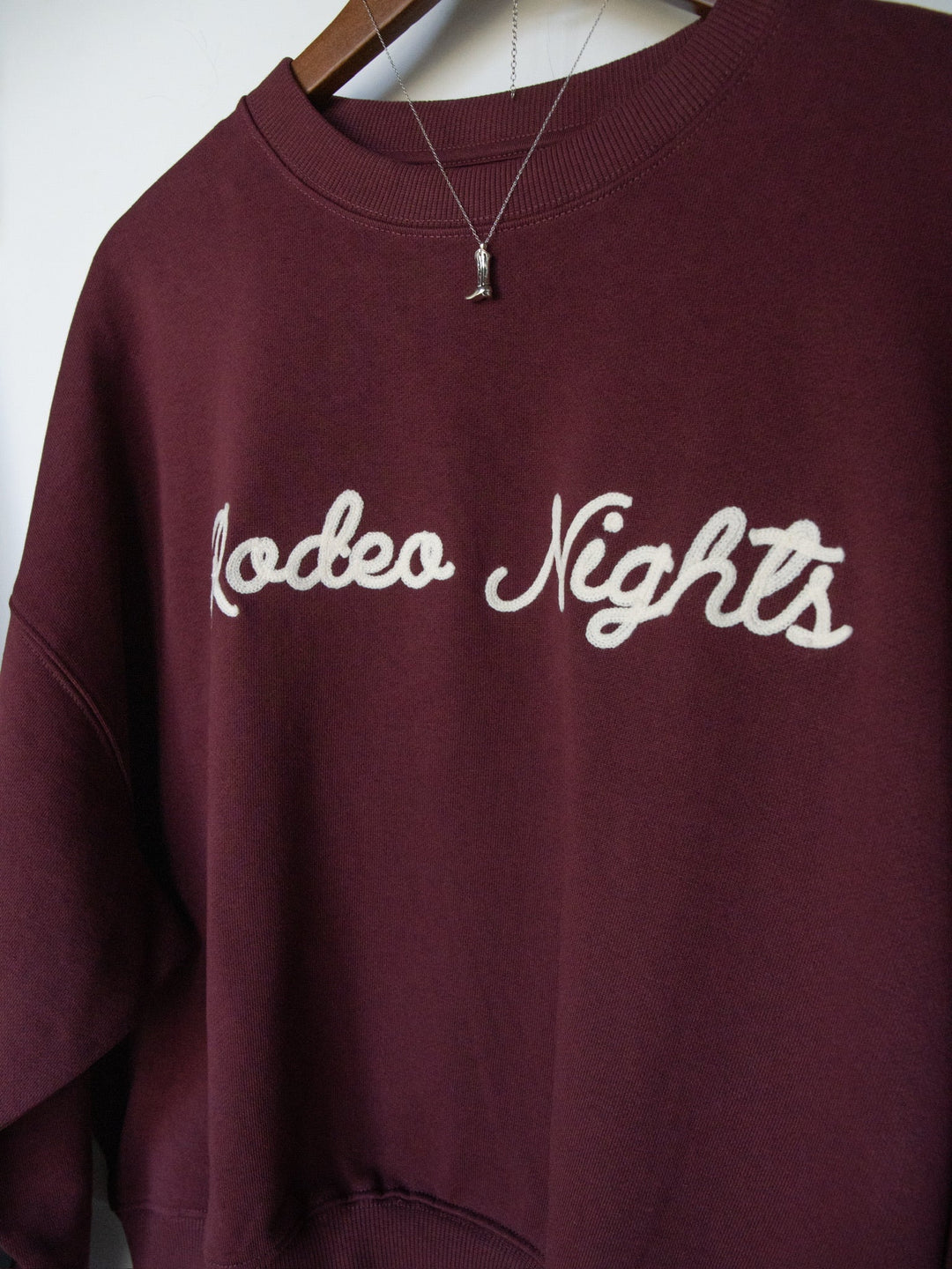 A burgundy long sleeve Rodeo Nights Graphic Pullover by Savannah Sevens western life{&}style, featuring “Rodeo Nights” in white script on the front. A silver necklace with a small pendant is layered over the pullover.