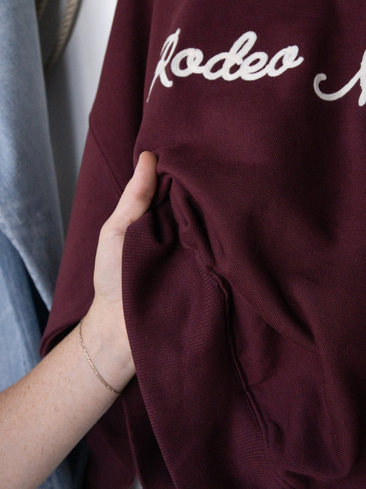A hand holds the Rodeo Nights Burgundy Graphic Pullover from Savannah Sevens western life{&}style, featuring white cursive "Rodeo" embroidery on a burgundy long sleeve sweatshirt. The person wears a delicate bracelet, highlighting the cozy style of this pullover.