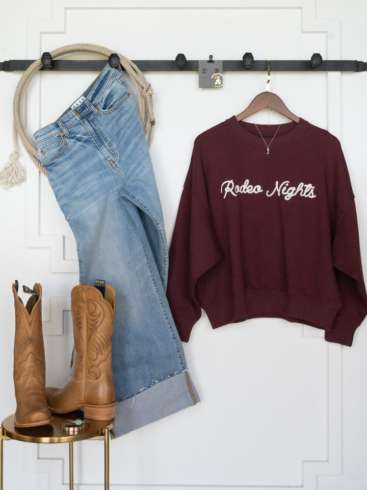 A pair of blue jeans and the Rodeo Nights Burgundy Graphic Pullover from Savannah Sevens western life{&}style hang on a wall. Brown cowboy boots rest on a stool below, creating the perfect rustic look inspired by the Lainey Wilson collection.