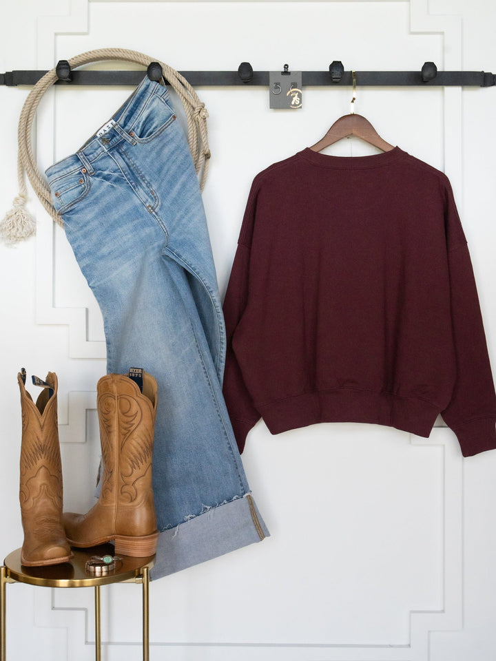 Displayed against a white wall, a pair of blue jeans and the Rodeo Nights Burgundy Graphic Pullover from Savannah Sevens western life{&}style hang side by side, while tan cowboy boots rest on a small round table below. Styled from the Lainey Wilson collection, this outfit pairs casual comfort with Western flair.