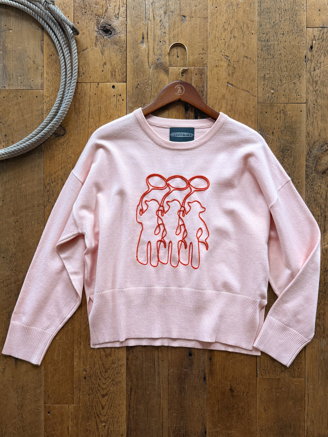 Roper Blush Western Embroidred Sweater in light pink with a relaxed fit, featuring a red embroidered line-art design of three figures on horseback on a wooden surface.