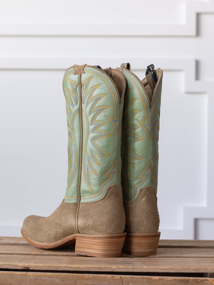 The Hyer Rose Hill Boot by Savannah Sevens western life{&}style features light green Western leather, tan suede bottoms, and wooden heels, displayed upright on wood against a white decorative wall.