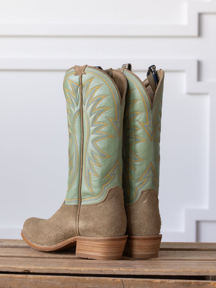 The Hyer Rose Hill Boot by Savannah Sevens western life{&}style features light green Western leather, tan suede bottoms, and wooden heels, displayed upright on wood against a white decorative wall.