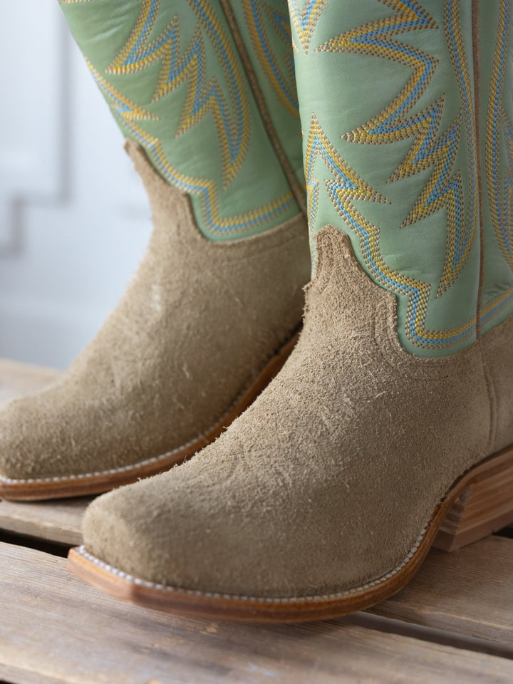 Close-up of The Hyer Rose Hill Boot by Savannah Sevens western life{&}style: tan suede Goodyear welt boots with pale green shafts and yellow-blue embroidered zigzags, resting on wood.