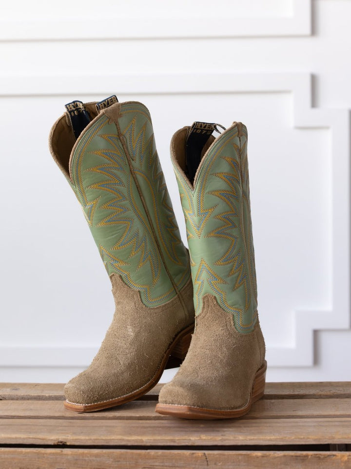 The Hyer Rose Hill Boot by Savannah Sevens western life{&}style features tan roughout leather, light green shafts, and yellow stitching, displayed side by side on a wooden surface against a white textured background.