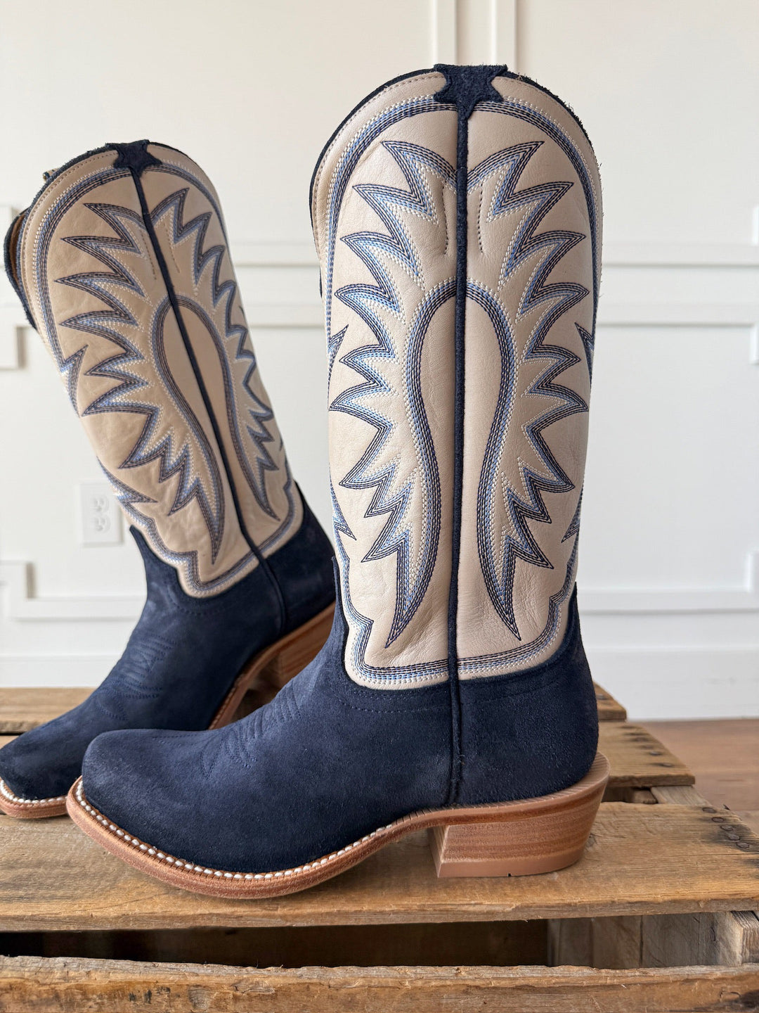 The Rose Hill Navy Western Leather Boot by Savannah Sevens western life{&}style is a pair of cutter toe boots featuring a navy blue suede foot and light tan leather shafts adorned with bold blue and gray stitched patterns. Displayed on a wooden surface indoors, these boots are crafted with a durable Goodyear leather welt and include a cushioned orthotic insert for all-day comfort. Boots by HYER. 