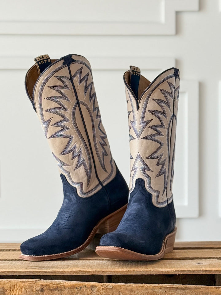 Introducing the Rose Hill Navy Western Leather Boot by Savannah Sevens western life{&}style: this standout pair features navy blue leather on the foot and heel, tan shafts accented by striking dark blue zigzag stitching, and brown soles with a sturdy Goodyear leather welt. Displayed on a wooden surface with a white paneled background. Boots by HYER. 