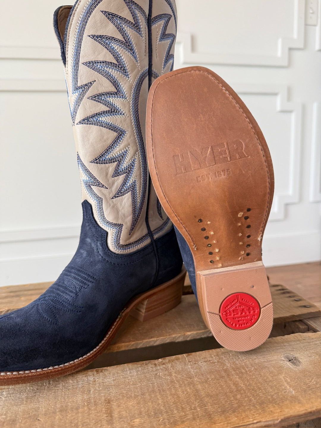 A close-up of the Savannah Sevens western life{&}style Rose Hill Navy Western Leather Boot on a wooden surface, featuring blue suede with white stitched shafts and a leather sole stamped with HYER EST. 1875 and a red logo near the heel. These Western cutter toe boots are crafted with a Goodyear leather welt for lasting durability.