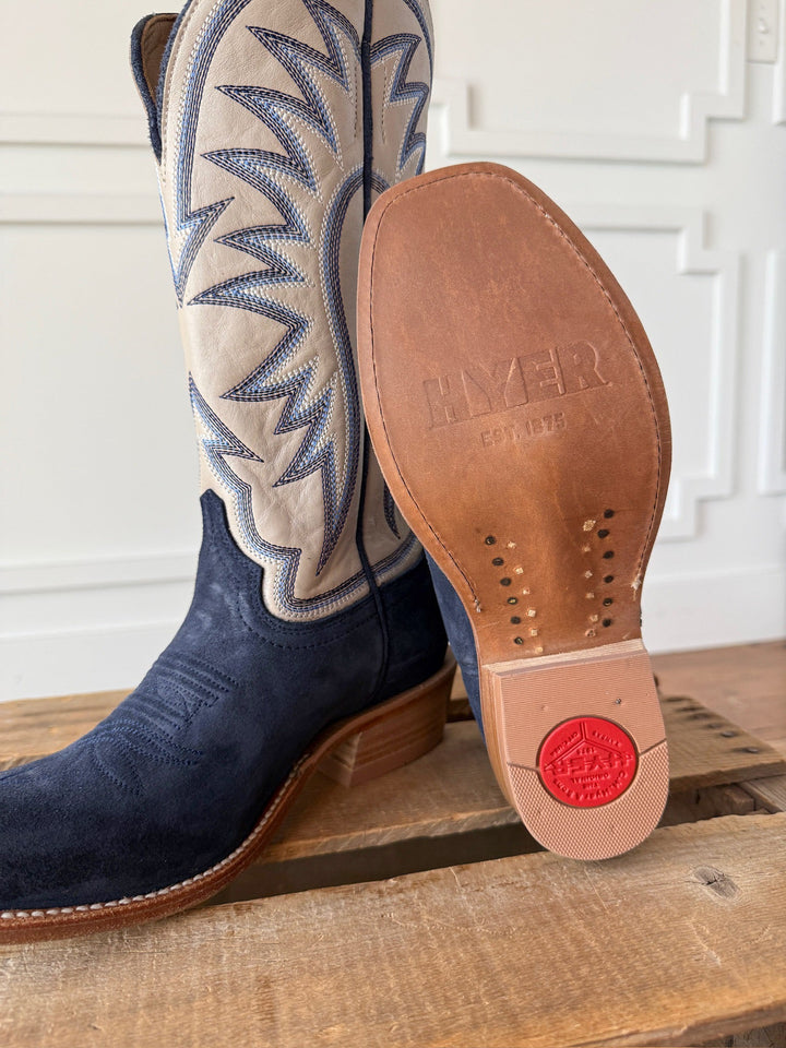 A close-up of the Savannah Sevens western life{&}style Rose Hill Navy Western Leather Boot on a wooden surface, featuring blue suede with white stitched shafts and a leather sole stamped with HYER EST. 1875 and a red logo near the heel. These Western cutter toe boots are crafted with a Goodyear leather welt for lasting durability.