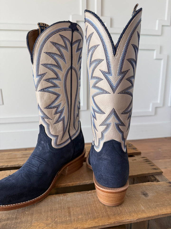 A pair of Rose Hill Navy Western Leather Boots by Savannah Sevens western life{&}style, showcasing navy blue suede lowers and cream tops adorned with detailed blue and silver stitching in a classic Western leather cutter toe silhouette, finished with a Goodyear leather welt, displayed on a wooden surface against a white paneled wall.