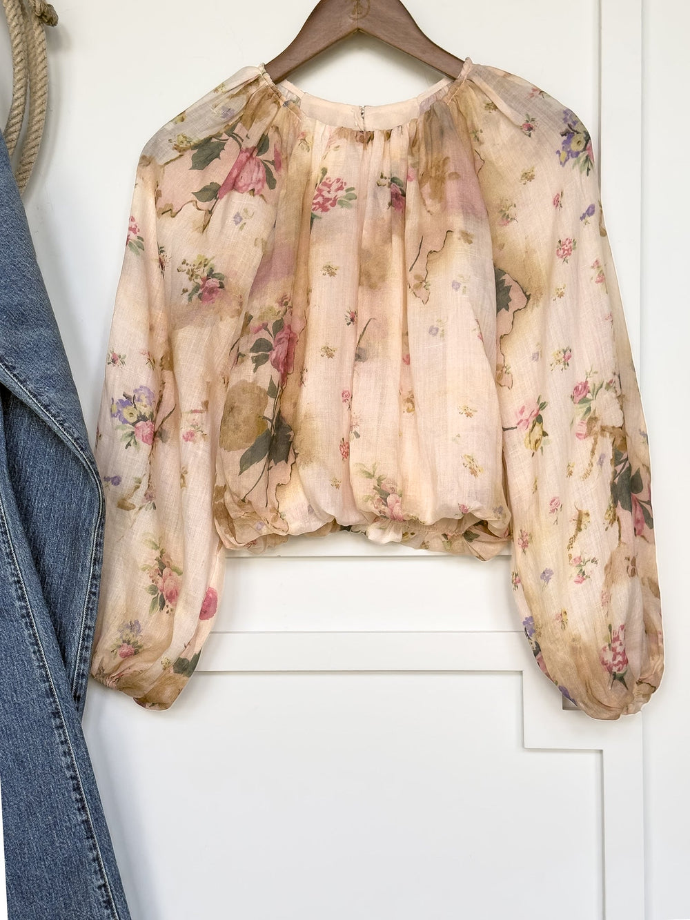 The Rosita Floral Bubble Hem Top by Savannah Sevens western life{&}style—a sheer, long-sleeve blouse with a pastel floral print, balloon sleeves, gathered neckline, and bubble hem—hangs on a wooden hanger next to a blue denim jacket. Its loose, billowy fit showcases its feminine style.