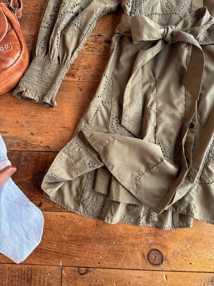 The Roswell Olive Eyelet Long Sleeve Dress by Savannah Sevens western life{&}style features a floral pattern, puffed sleeves, and a waist tie. It rests on rustic wood beside part of a tan purse and a white fabric edge in the lower left corner.