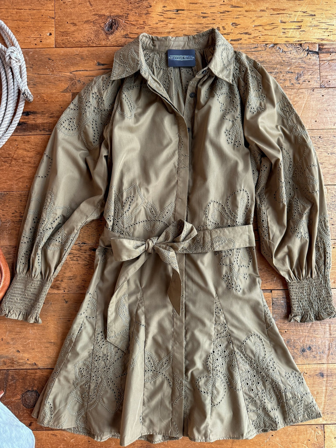 The Roswell Olive Eyelet Long Sleeve Dress by Savannah Sevens western life{&}style features floral eyelet embroidery, a collared neckline, long sleeves with smocked cuffs, and a matching waist tie, displayed on a rustic wooden surface.
