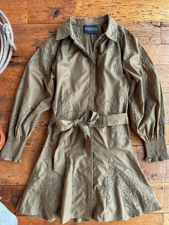 The Roswell Olive Eyelet Long Sleeve Dress by Savannah Sevens western life{&}style features floral eyelet embroidery, a collared neckline, long sleeves with smocked cuffs, and a matching waist tie, displayed on a rustic wooden surface.
