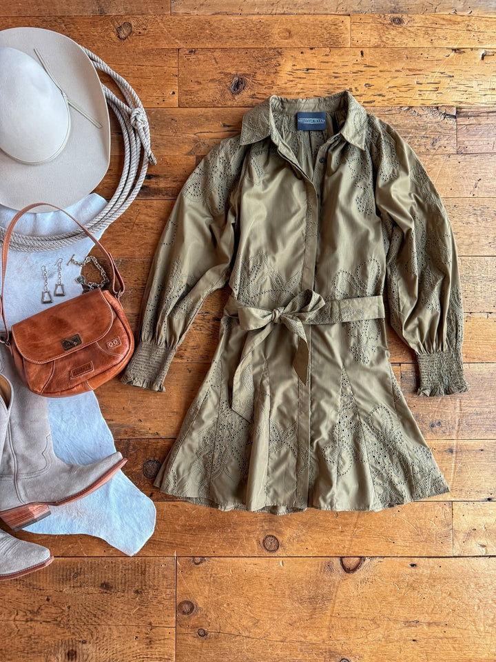 The Roswell Olive Eyelet Long Sleeve Dress by Savannah Sevens western life{&}style is laid out on a wooden floor, paired with a beige wide-brim hat, brown leather purse, silver jewelry, and light suede boots.