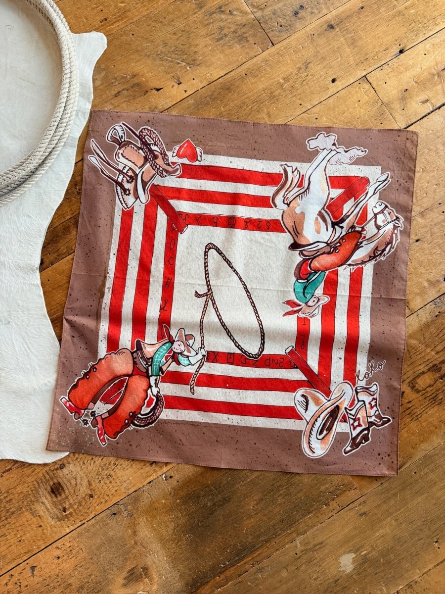 The Round Up Neck Scarf by Savannah Sevens western life{&}style, featuring red and white stripes with cowboy illustrations in each corner, is laid out on a wooden floor next to a white rope and cloth.