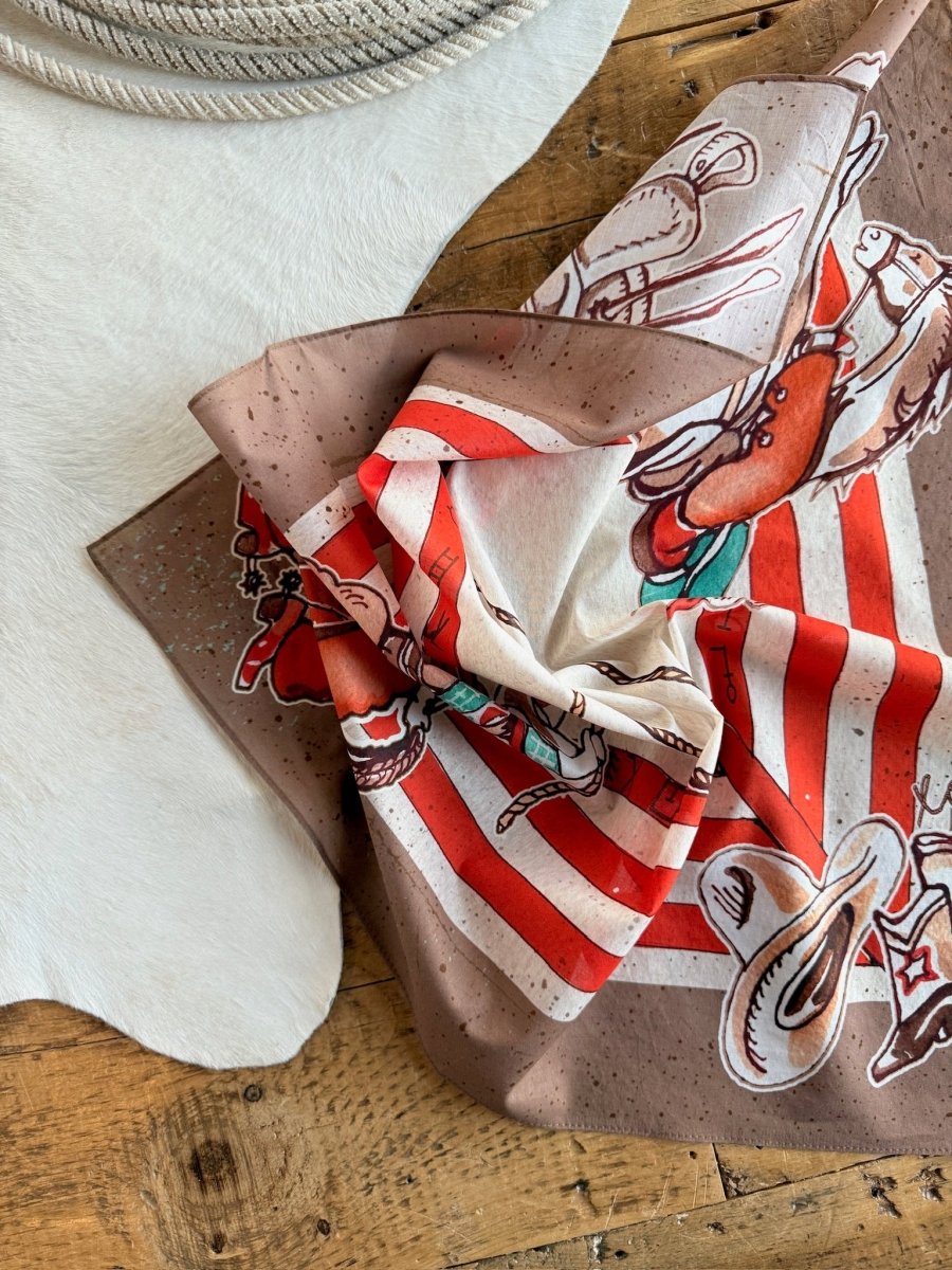 The Round Up Neck Scarf by Savannah Sevens western life{&}style, featuring red and white stripes with retro rodeo designs, is crumpled on a wooden floor beside a white cowhide rug and a coiled rope.