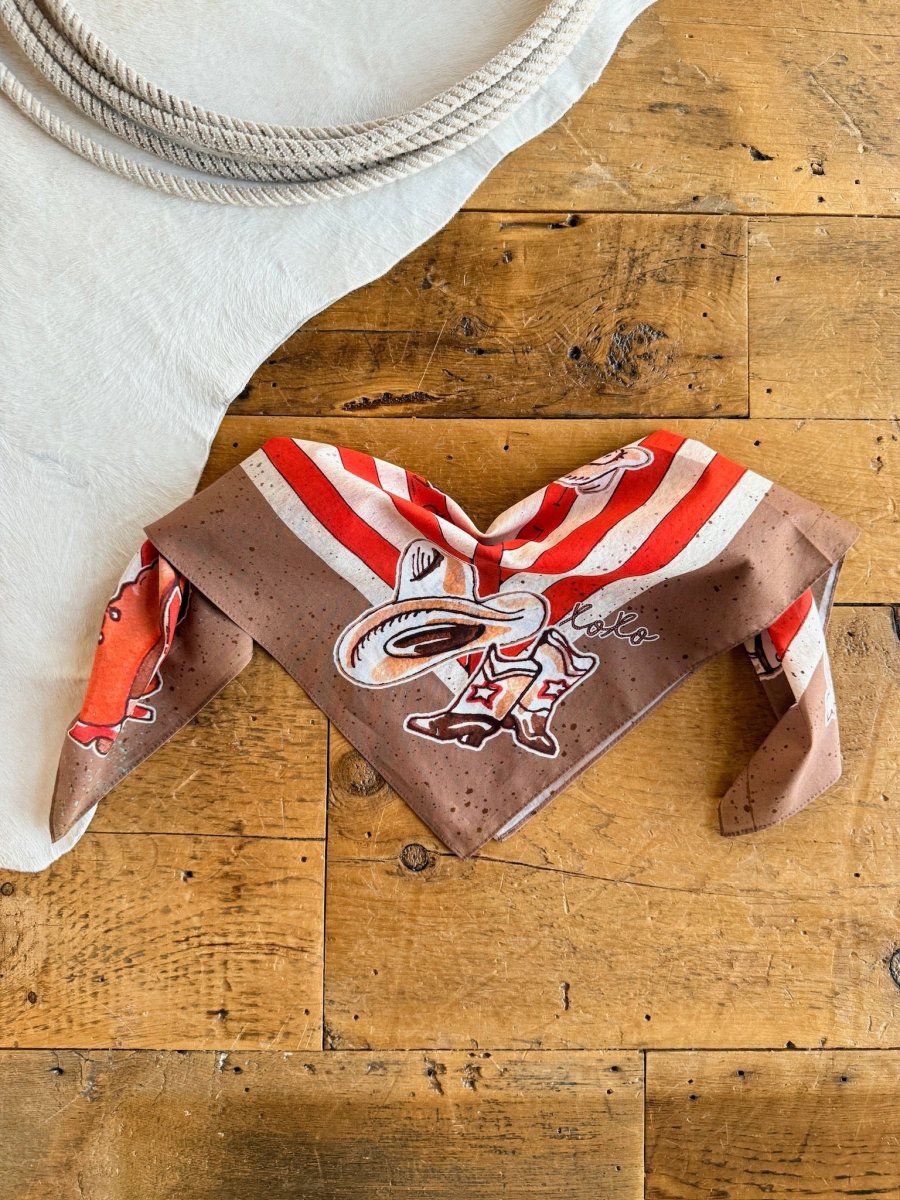 The Round Up Neck Scarf by Savannah Sevens western life{&}style, featuring brown and red retro rodeo stripes, is folded on a rustic wooden surface near coiled rope and white fabric.