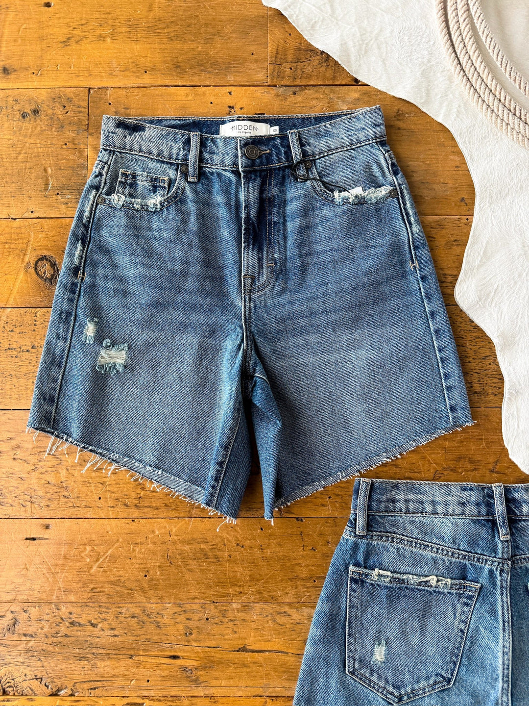 The Rowan Shorts by Savannah Sevens western life{&}style are vintage denim shorts with a raw hem and distressed details on the front and back, shown laid flat beside a white fabric on a wooden surface.