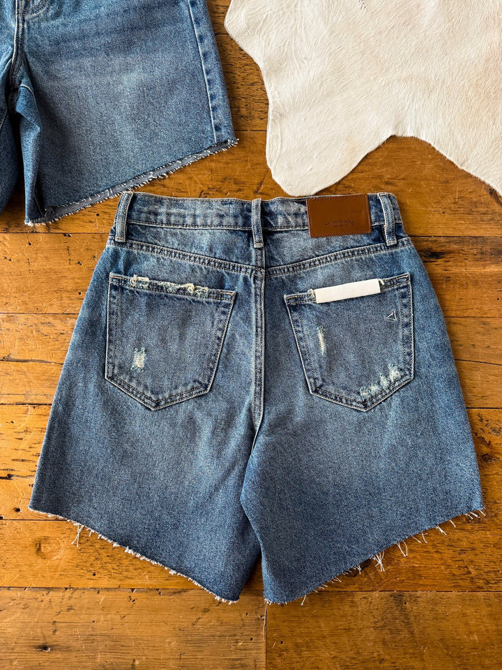 The Rowan Shorts by Savannah Sevens western life{&}style are shown from the back on a wooden floor with frayed hems and a white card in the right pocket. Another pair of shorts and part of a white rug are also visible.